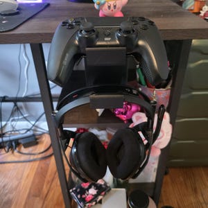 Puede incluir: Un mando de juegos negro y unos auriculares se muestran en un soporte de metal negro. El mando está en la parte superior y los auriculares cuelgan debajo. Los auriculares tienen auriculares grandes y negros y un micrófono. El soporte está en un estante marrón oscuro.