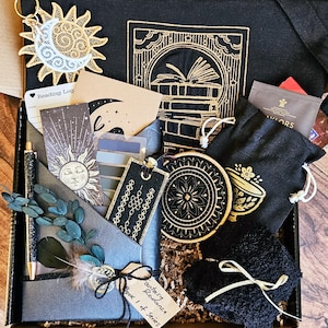 May include: A black box filled with a variety of bookish items, including a reading log, a bookmark, a teacup coaster, a black tote bag with a gold embroidered book design, and a small black bag with a gold embroidered teacup design. The box is filled with brown packing paper and the text "Book 1 of Series" is visible on a tag.