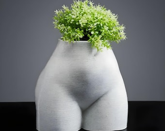 Könnte beinhalten: Eine weiße Keramikvase in Form eines Torsos mit strukturierter Oberfläche. Die Vase ist mit einem Büschel kleiner, hellgrüner Kunstpflanzen gefüllt. Die Vase steht auf einer schwarzen Oberfläche.