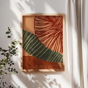Puede incluir: Impresión artística abstracta enmarcada, con una gran forma de hoja roja estilizada y una forma verde, rayada y curvada sobre un fondo melocotón. La obra de arte está en un marco de madera clara, creando una estética moderna y minimalista.