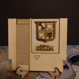 May include: A wooden cutout of a Nintendo Entertainment System (NES) game cartridge featuring the iconic "The Legend of Zelda" logo and a shield with a key, hearts, and a lion. The cutout is on a stand.