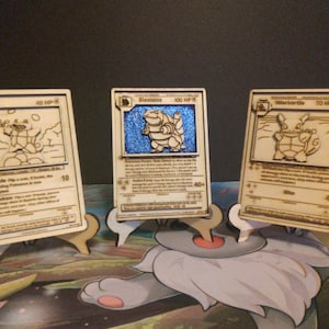 Może przedstawiać: Trzy drewniane karty Pokemon z podstawkami. Karty przedstawiają Squirtle, Blastoise i Wartortle. Karty są wycięte laserowo i mają błyszczące niebieskie tło.