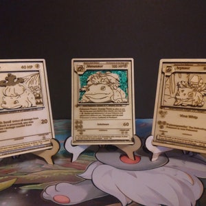May include: Three wooden Pokemon cards featuring Bulbasaur, Ivysaur, and Venusaur. The cards are displayed on small wooden stands. The cards are laser cut and have a detailed design.