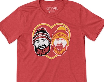Può includere: Maglietta rossa con una grafica a forma di cuore che presenta i volti di due giocatori di football, Jason e Travis, che indossano cappelli rossi e gialli con "KC" su di essi. Il testo "CITYSOUL" è visibile sull'etichetta del collo della maglietta.