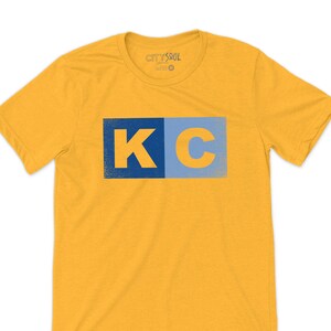 May include: A yellow t-shirt with a blue and light blue "KC" graphic on the front. The shirt has a crew neck and short sleeves. The brand name "CITYSOUL" is visible on the neck label.