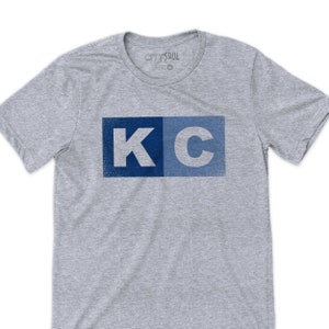 May include: A gray t-shirt with a blue and light blue "KC" graphic printed on the front. The graphic has a distressed look.