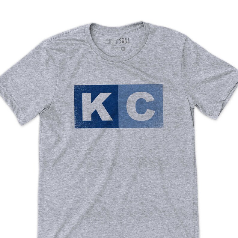 May include: A gray t-shirt with a blue and light blue "KC" graphic printed on the front. The graphic has a distressed look.