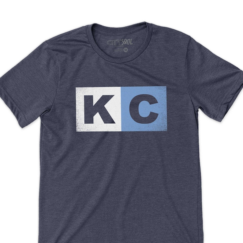 May include: A navy blue t-shirt with a white and blue graphic of the letters "KC" in a vintage style.