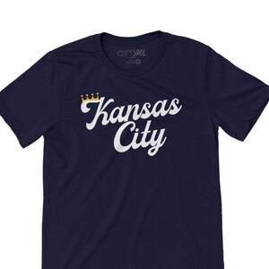 Puede incluir: Una camiseta azul marino con un gr&aacute;fico blanco de las palabras "Kansas City" en una fuente retro con una corona dorada sobre la palabra "Kansas".