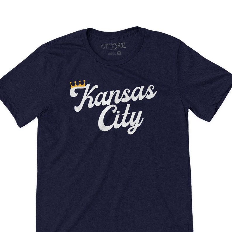 Puede incluir: Una camiseta azul marino con un gr&aacute;fico blanco de las palabras "Kansas City" en una fuente retro con una corona dorada sobre la palabra "Kansas".