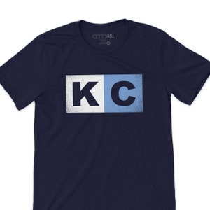 May include: A navy blue t-shirt with a white and light blue graphic of the letters "KC" in a distressed style.