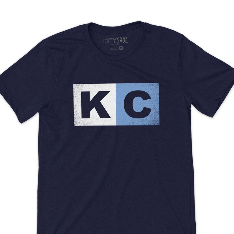May include: A navy blue t-shirt with a white and light blue graphic of the letters "KC" in a distressed style.