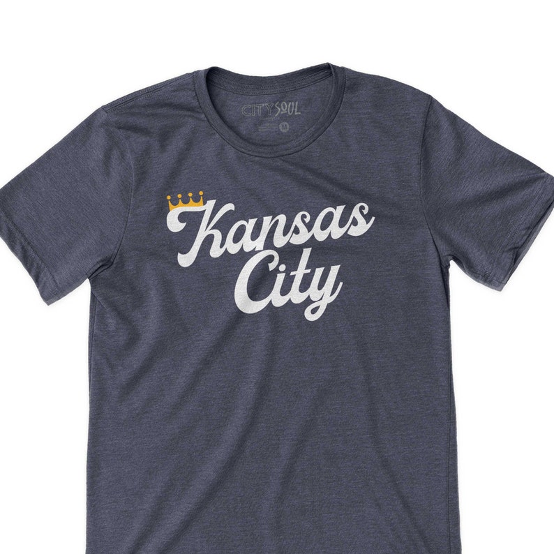 Puede incluir: Una camiseta azul marino con un gr&aacute;fico blanco que dice "Kansas City" con una corona dorada sobre la palabra "Kansas".