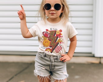 May include: A young girl wearing a white t-shirt with a graphic design featuring a lightning bolt, a football, a smiley face, and the text "KC Rocks" and "Kansas City". She is also wearing denim shorts and sunglasses.