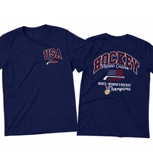 May include: Navy blue t-shirt with "USA" and a hockey stick graphic on the front. The back features "HOCKEY Milano Cortina" text above a flag graphic, and "MEN'S WOMEN'S HOCKEY Champions" text with a gold medal.