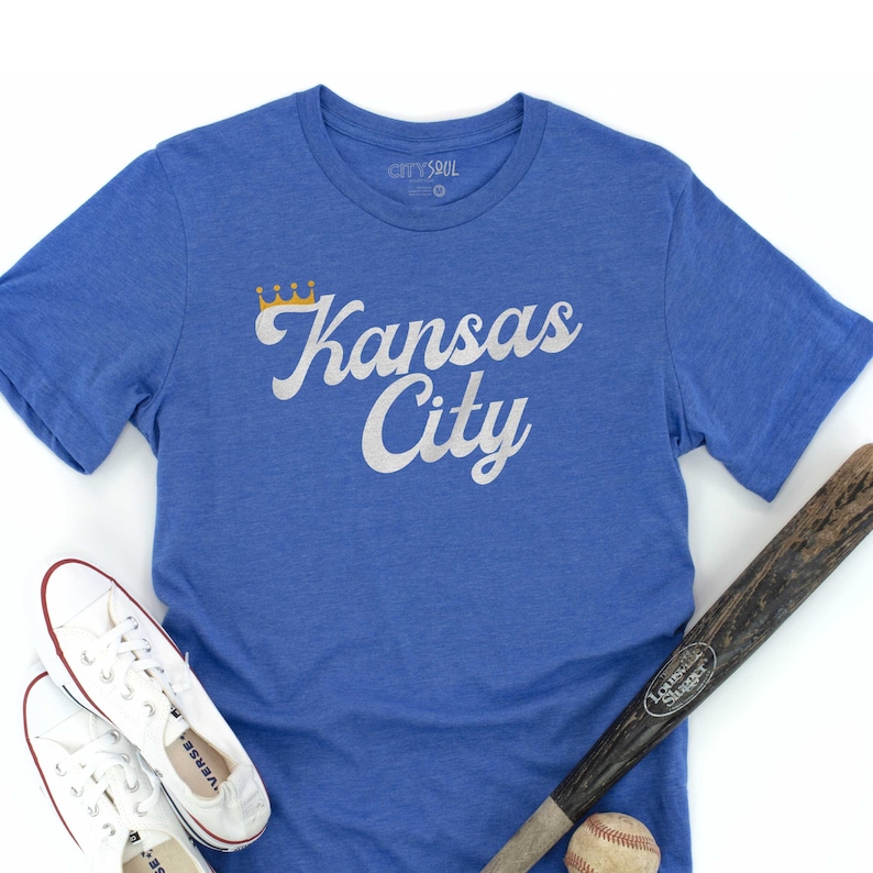 Puede incluir: Una camiseta azul real con un gr&aacute;fico blanco que dice "Kansas City" con una corona dorada sobre la palabra "Kansas".