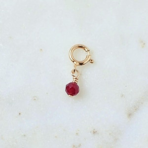 May include: A gold spring ring clasp charm featuring a small, faceted, deep red gemstone bead suspended from a gold wire loop. The circular clasp mechanism is designed for attachment to jewelry chains, showcasing a polished metallic finish.
