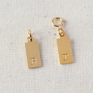 May include: Two gold-colored rectangular charms with engraved letters. One charm has the letter "R" and the other has the letter "T". Both charms have a small loop at the top for attaching to a necklace or bracelet.