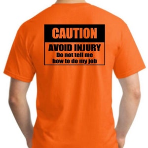 Pu&ograve; includere: Maglietta arancione con un cartello di avvertenza nero che dice "CAUTION AVOID INJURY Do not tell me how to do my job"