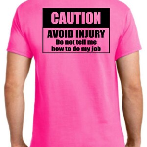 Pu&ograve; includere: Una maglietta rosa con un cartello di attenzione nero che dice "CAUTION AVOID INJURY Do not tell me how to do my job".