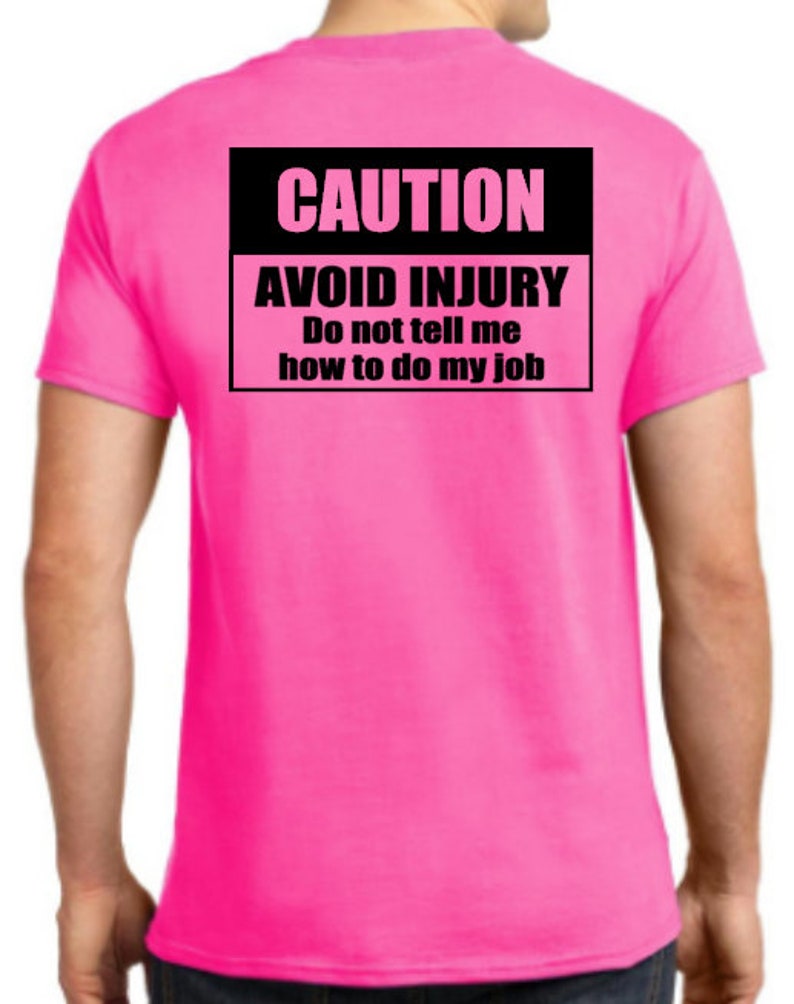 Pu&ograve; includere: Una maglietta rosa con un cartello di attenzione nero che dice "CAUTION AVOID INJURY Do not tell me how to do my job".