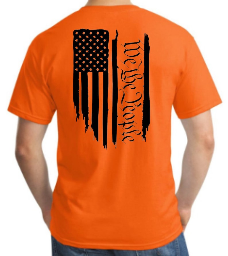 Puede incluir: Camiseta naranja con una bandera estadounidense negra desgastada y el texto "We the People" impreso en la espalda.