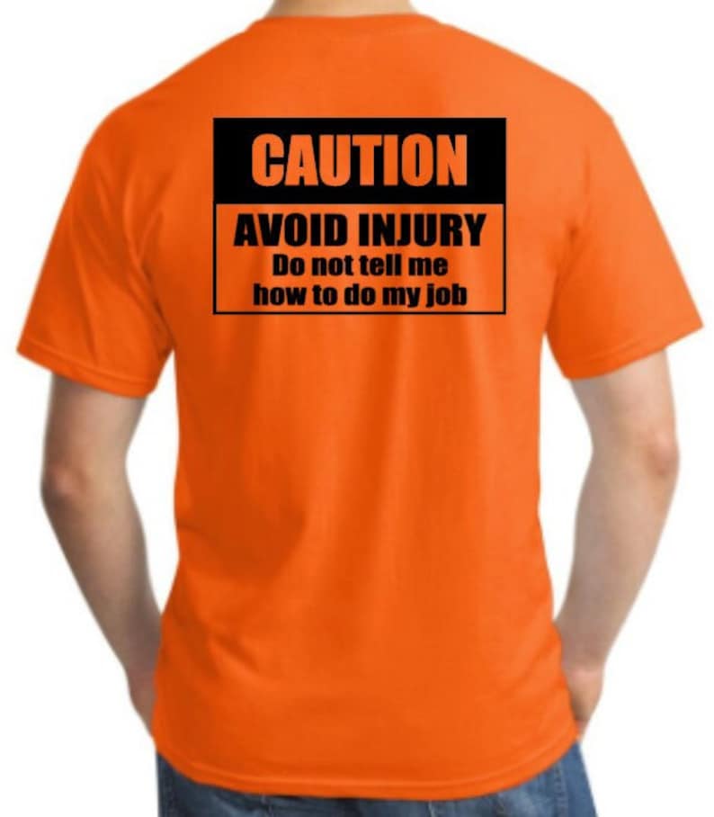 Pu&ograve; includere: Maglietta arancione con un cartello di avvertenza nero che dice "CAUTION AVOID INJURY Do not tell me how to do my job"