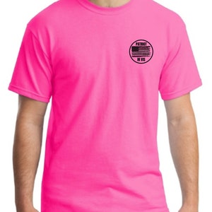 Pu&ograve; includere: Una maglietta rosa brillante con una toppa circolare nera sul petto sinistro. La toppa presenta una bandiera americana in bianco e nero con le parole "PATRIOT HI VIS" scritte sotto.