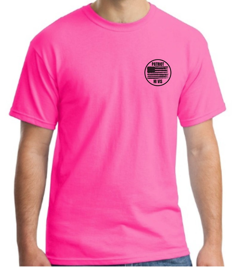 Pu&ograve; includere: Una maglietta rosa brillante con una toppa circolare nera sul petto sinistro. La toppa presenta una bandiera americana in bianco e nero con le parole "PATRIOT HI VIS" scritte sotto.