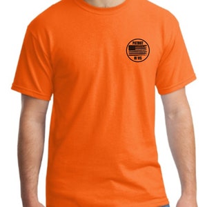 Pu&ograve; includere: T-shirt arancione a maniche corte con una toppa circolare nera sul petto sinistro. La toppa presenta una bandiera americana in bianco e nero e il testo "PATRIOT HI VIS".