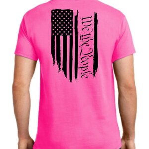 Puede incluir: Una camiseta rosa con una bandera estadounidense negra desgastada y el texto "We the People" impreso en la espalda.