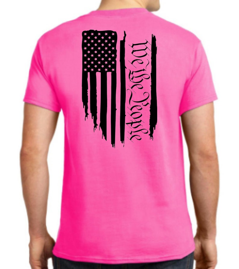 Puede incluir: Una camiseta rosa con una bandera estadounidense negra desgastada y el texto "We the People" impreso en la espalda.