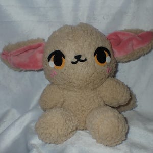 May include: A plush toy shaped like a small animal with large, pink ears, brown fur, and black eyes. The toy has a cute, cartoon-like face with a small, black nose and a pink blush on its cheeks.