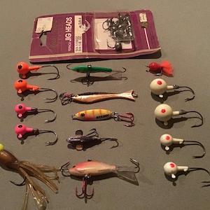 Könnte beinhalten: Eine Sammlung von Angelködern in verschiedenen Farben und Designs, darunter Orange, Grün, Rosa und Weiß. Einige Köder haben eine fischähnliche Form, während andere einen runden Kopf mit einem Haken aufweisen. Eine Packung Jig-Köpfe ist ebenfalls sichtbar.
