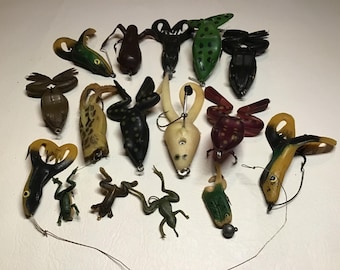 May include: A collection of vintage fishing lures in various frog designs. The lures are in shades of green, yellow, brown, and black, with detailed features and attached hooks. Some have spots or stripes, and some have metal weights.