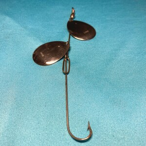 May include: A silver-colored fishing lure with two oval-shaped blades and a single hook. The blades are attached to a wire frame, and the hook is at the bottom. The blades have text on them. The lure is designed to attract fish.