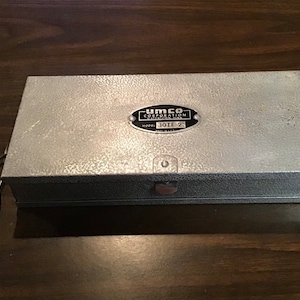 May include: A rectangular, silver-colored metal box with a textured surface. The box has a small metal handle and a label that reads "UMCO CORPORATION" and "MODEL 101F-2". The box is closed and sits on a dark wooden surface.