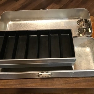 Può includere: Fornello da campeggio vintage in metallo argentato, aperto per rivelare un vassoio in plastica nera con sei scomparti. Il fornello ha un coperchio incernierato e un piccolo gruppo bruciatore. Su una superficie di legno.