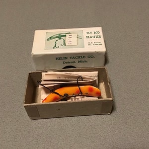 Puede incluir: Señuelo Fly-Rod Flatfish de Helin Tackle Co. vintage en su caja original. El señuelo es naranja y blanco con anzuelos negros. La caja es blanca con un dibujo del señuelo y el texto "FLY-ROD FLATFISH". La caja también tiene el texto "HELIN TACKLE CO. Detroit, Mich."