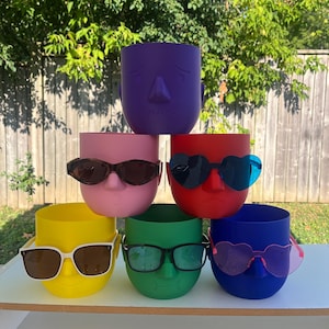 May include: A stack of six brightly coloured, face-shaped planters. The planters are in shades of purple, pink, red, yellow, green, and blue. Each planter has a face design and is adorned with sunglasses. The planters are stacked on a white surface.