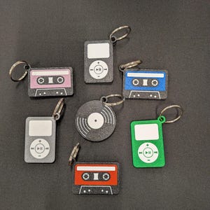 May include: A collection of keychains featuring retro designs. The keychains include a pink cassette tape, a blue cassette tape, a red cassette tape, two gray iPods, a green iPod, and a vinyl record. Each keychain has a silver keyring.