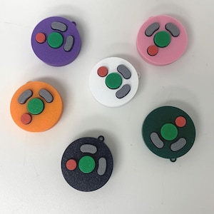 May include: A selection of colourful fidget toys in shades of purple, pink, orange, white, navy, and green. Each toy has a circular design with a green centre button, grey side buttons, and a red accent button.