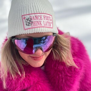 May include: White knitted beanie with a pink patch that reads "Dance First Think Later". The beanie is being worn by a person wearing pink fur and mirrored sunglasses.