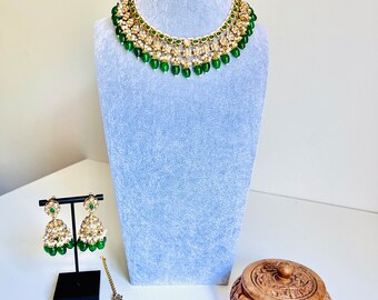May include: A gold and green Indian bridal necklace set with matching earrings and a tikka. The necklace has multiple layers of gold beads and green stones, and the earrings and tikka have a similar design.