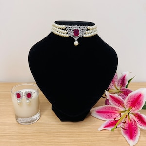 May include: A three-strand pearl choker necklace with a silver pendant featuring a red gemstone surrounded by clear rhinestones. Matching earrings with red gemstones and pearl drops.