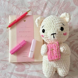 Puede incluir: Un oso de peluche blanco tejido con un chal rosa y un pequeño libro rosa. Un bolígrafo rosa y dos pintalabios están junto a un libro con el texto "we love because he first loved us". El fondo es un estampado floral.