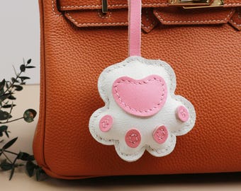 May include: A brown leather handbag with a white and pink leather paw print charm attached to it.
