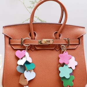 May include: A brown leather handbag with gold hardware and two charms. One charm is a cluster of colorful hearts and the other is a cluster of colorful stars.