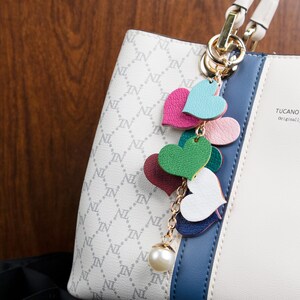 May include: A white handbag with a blue stripe and a gold chain with a pearl charm and a series of colorful leather hearts. The bag has a repeating pattern of "IN" and "NL". The bag has a label that says "TUCANO Original".
