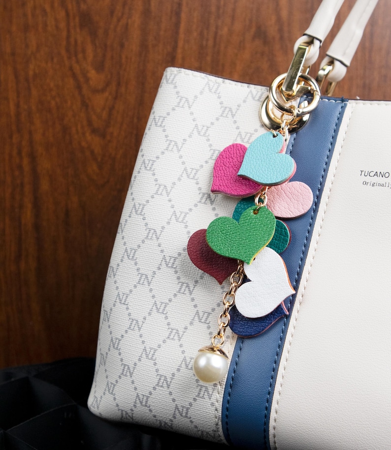 May include: A white handbag with a blue stripe and a gold chain with a pearl charm and a series of colorful leather hearts. The bag has a repeating pattern of "IN" and "NL". The bag has a label that says "TUCANO Original".